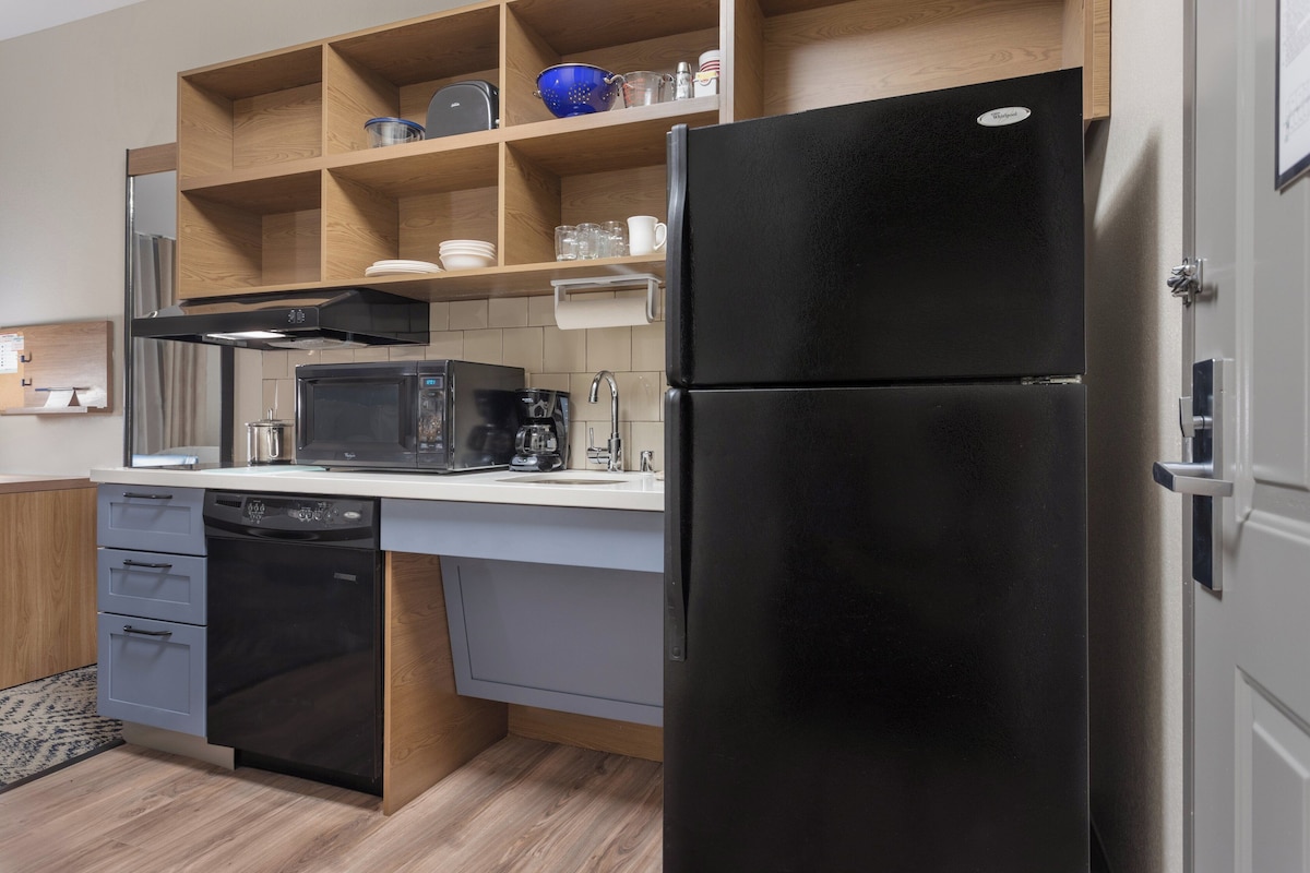 A modern kitchen with a black refrigerator, a microwave, a dishwasher, and wooden shelves above the counter.
