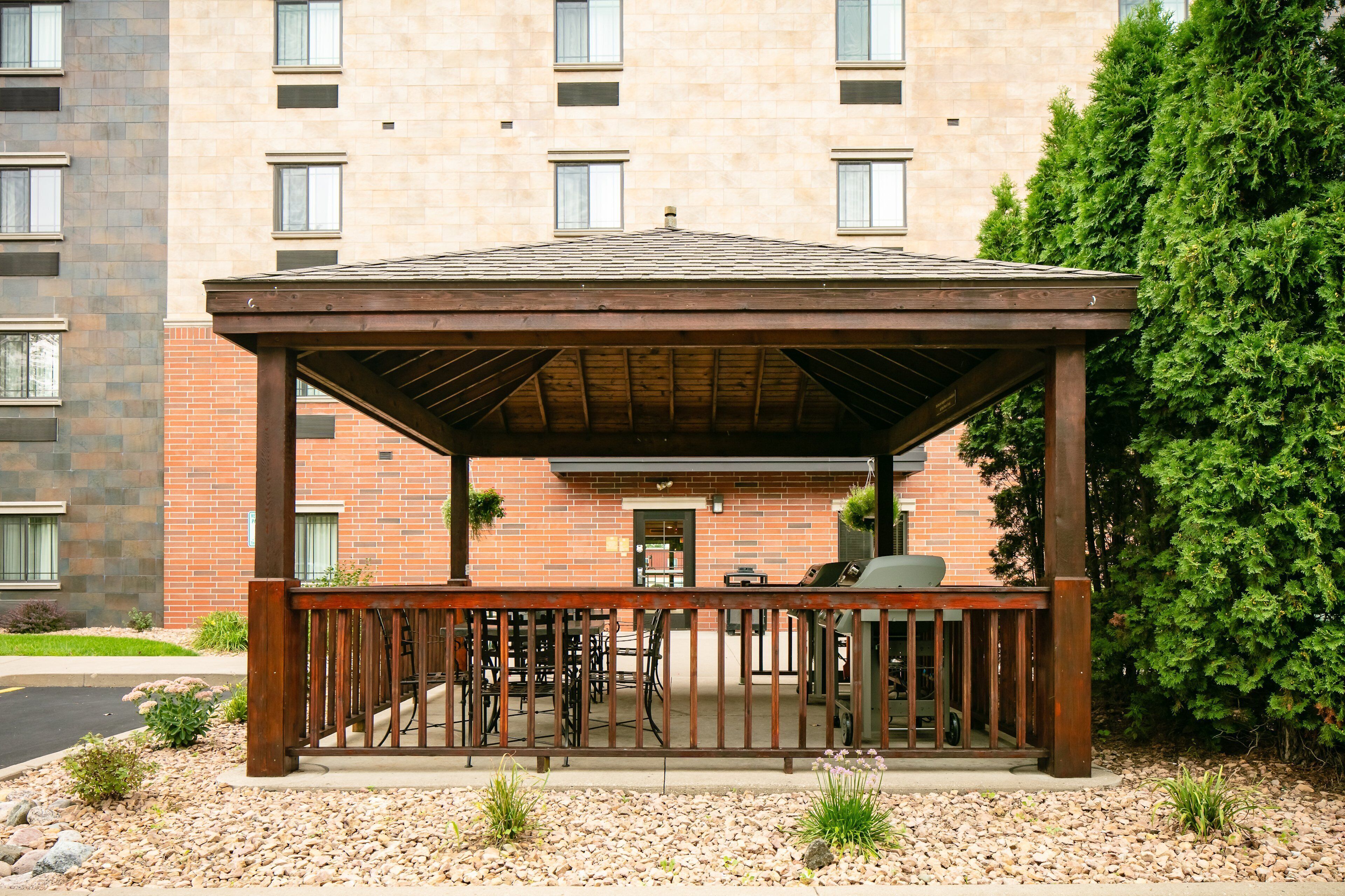 A covered outdoor seating area with a wooden deck and metal railing, surrounded by a gravel path and small plants.