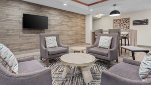 Un hall d’entr ée moderne avec des fauteuils à revêtement gris, une table basse ronde en bois et un téléviseur à écran plat fixé à un mur en bois.