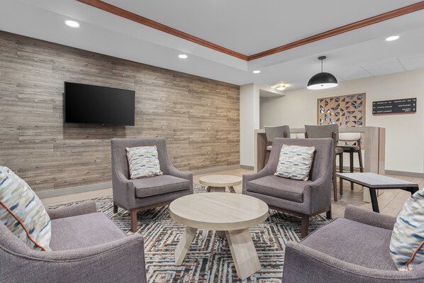 Un hall d’entrée moderne avec des fauteuils à revêtement gris, une table basse ronde en bois et un téléviseur à écran plat fixé à un mur en bois.