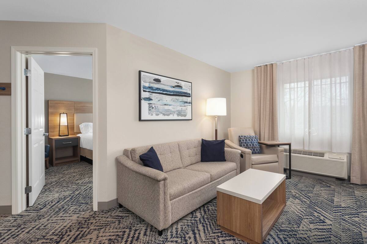 Une chambre d’hôtel avec un canapé beige, une table basse blanche, une œuvre d’art encadrée au mur, une lampe, une chaise et une grande fenêtre avec des rideaux.