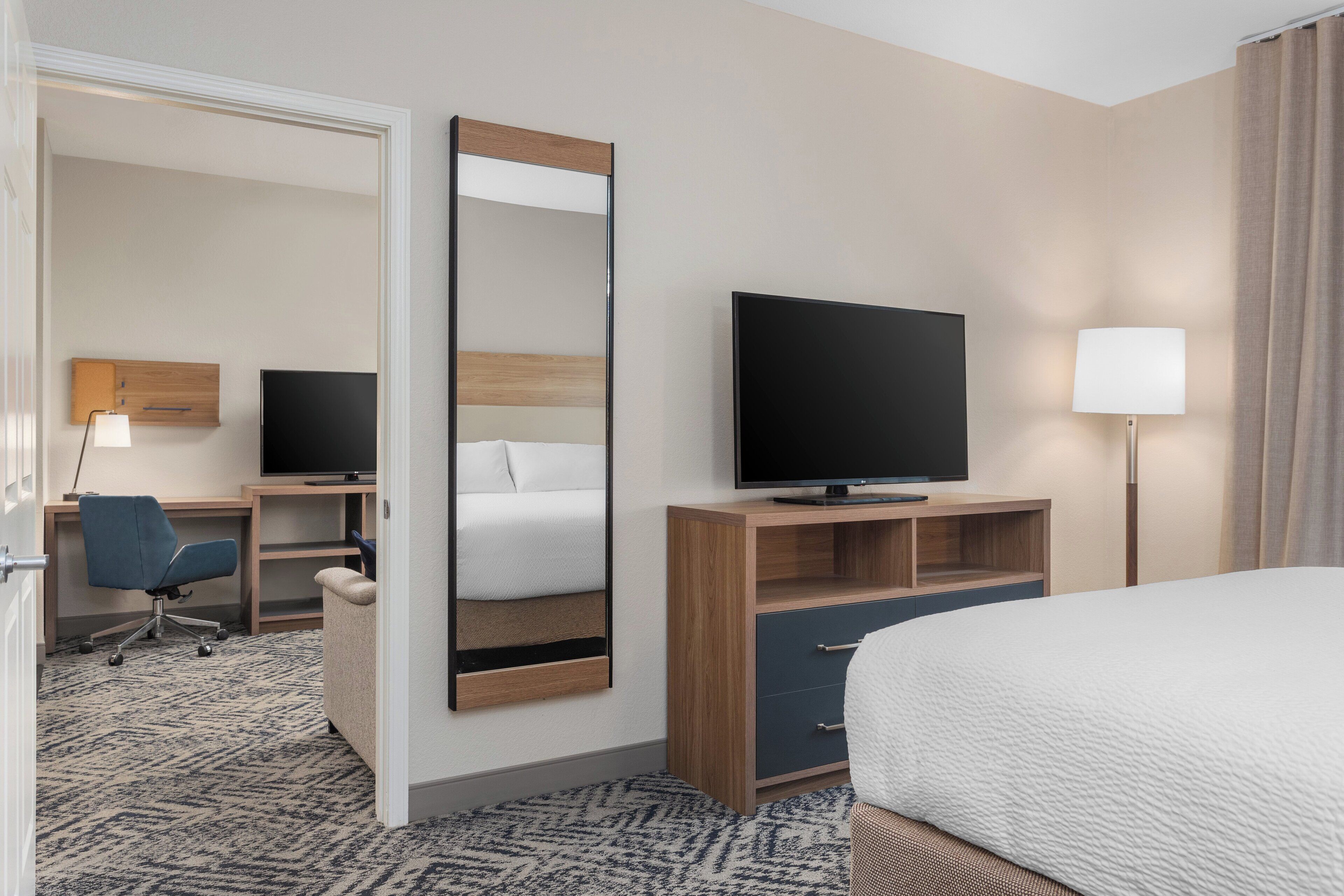 A hotel room with a bed, a TV, a desk with a chair, a mirror, and a lamp.