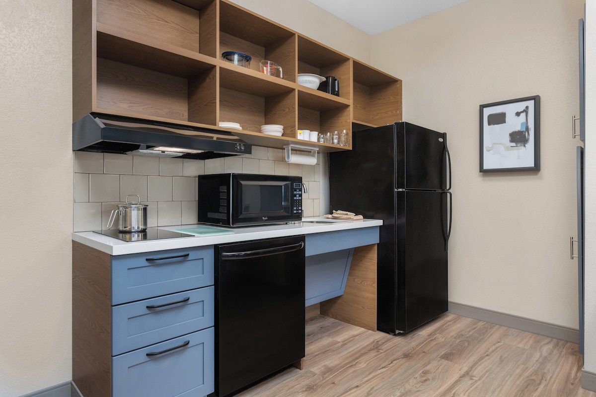 A compact kitchen with a microwave, dishwasher, and black refrigerator. There are wooden shelves above the counter holding dishes and glasses.