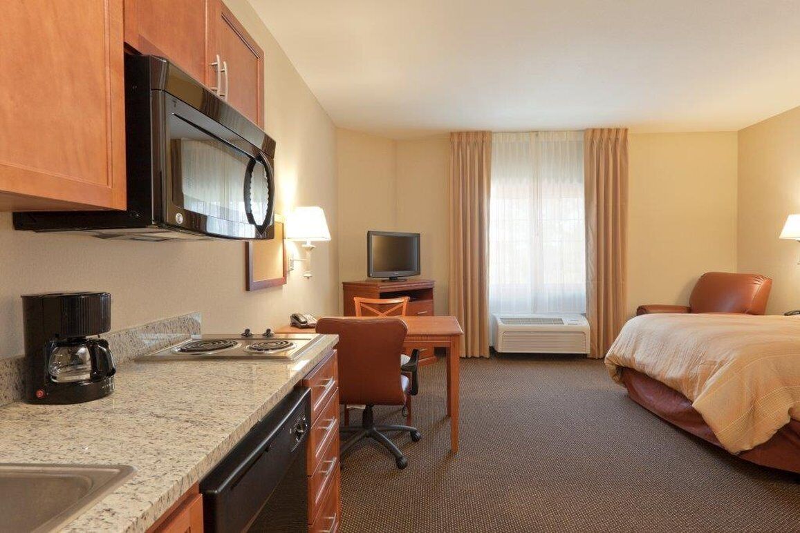 A hotel room with a kitchenette, a bed, a desk with a chair, a television, and a window with curtains.
