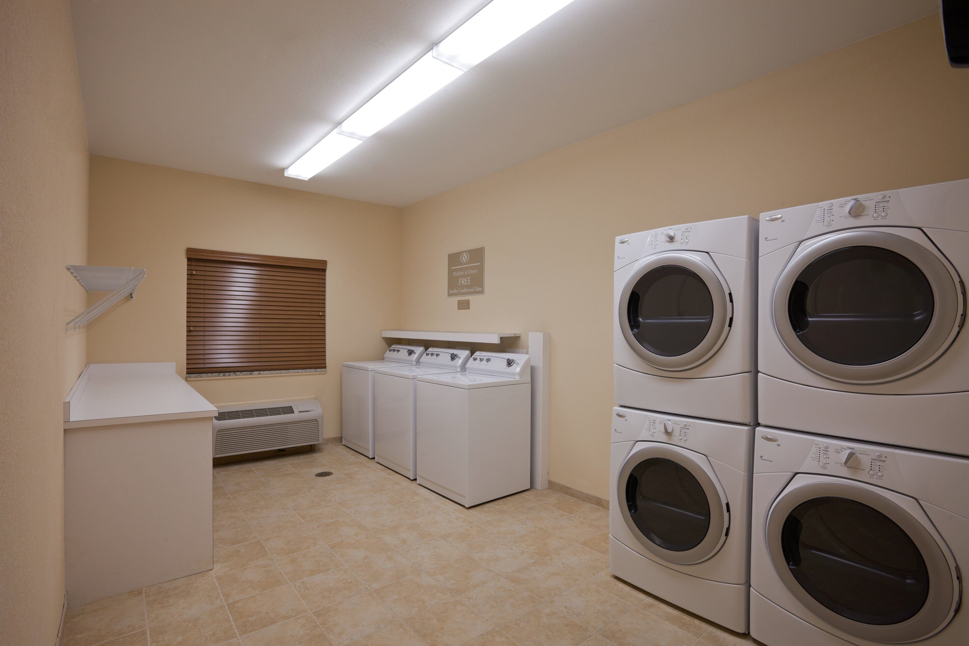 A laundry room with four stacked front-loading washers and dryers, a wall-mounted heater, and a window with blinds.