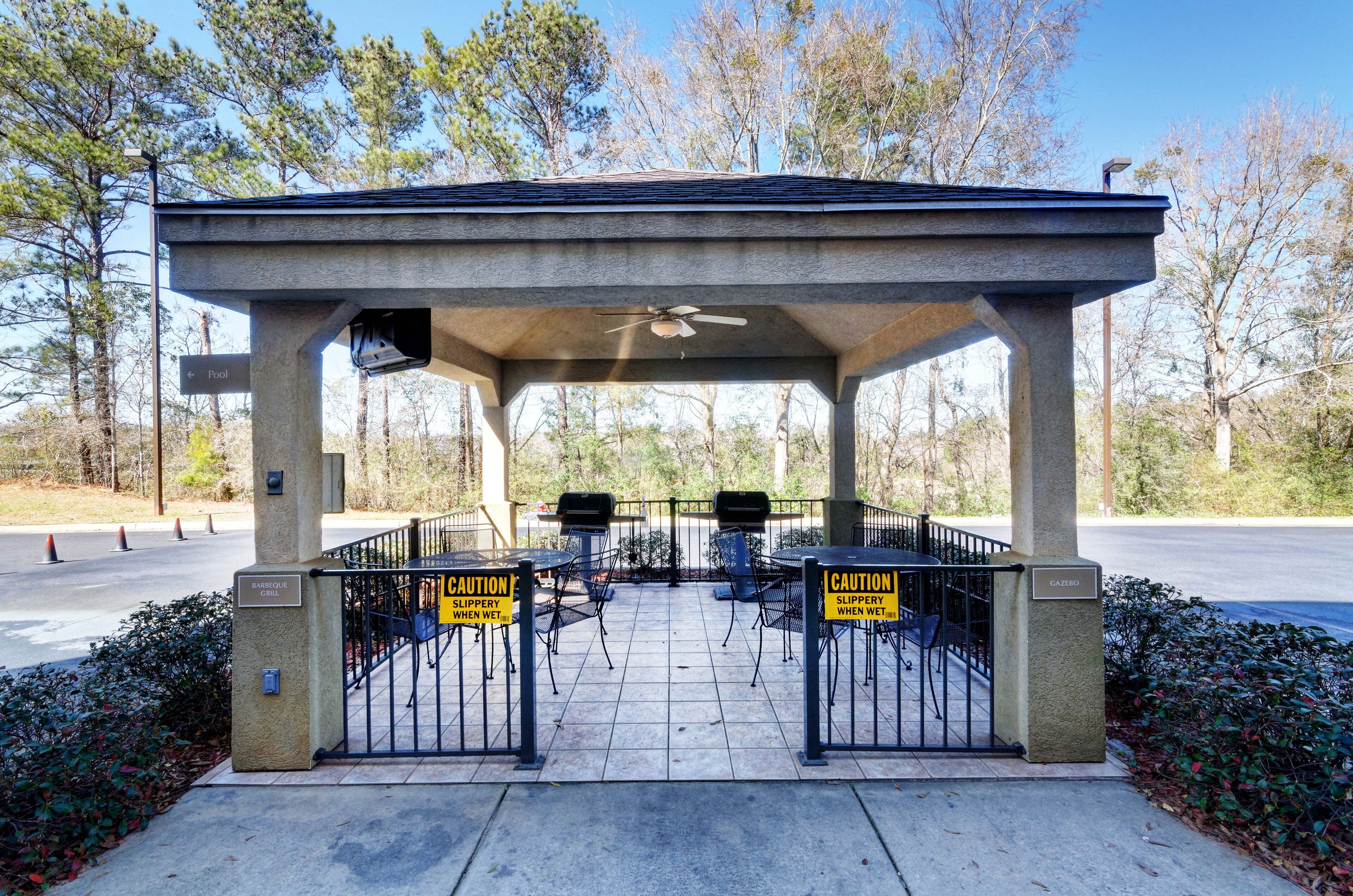 Un espacio de estar al aire libre techado con una parrilla, un letrero de precaución por superficies resbaladizas cuando están mojadas, y una reja que da a un estacionamiento.