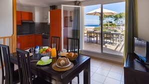 A dining area with a table set for breakfast, a kitchen in the background, and a balcony with outdoor seating and a view of the sea.
