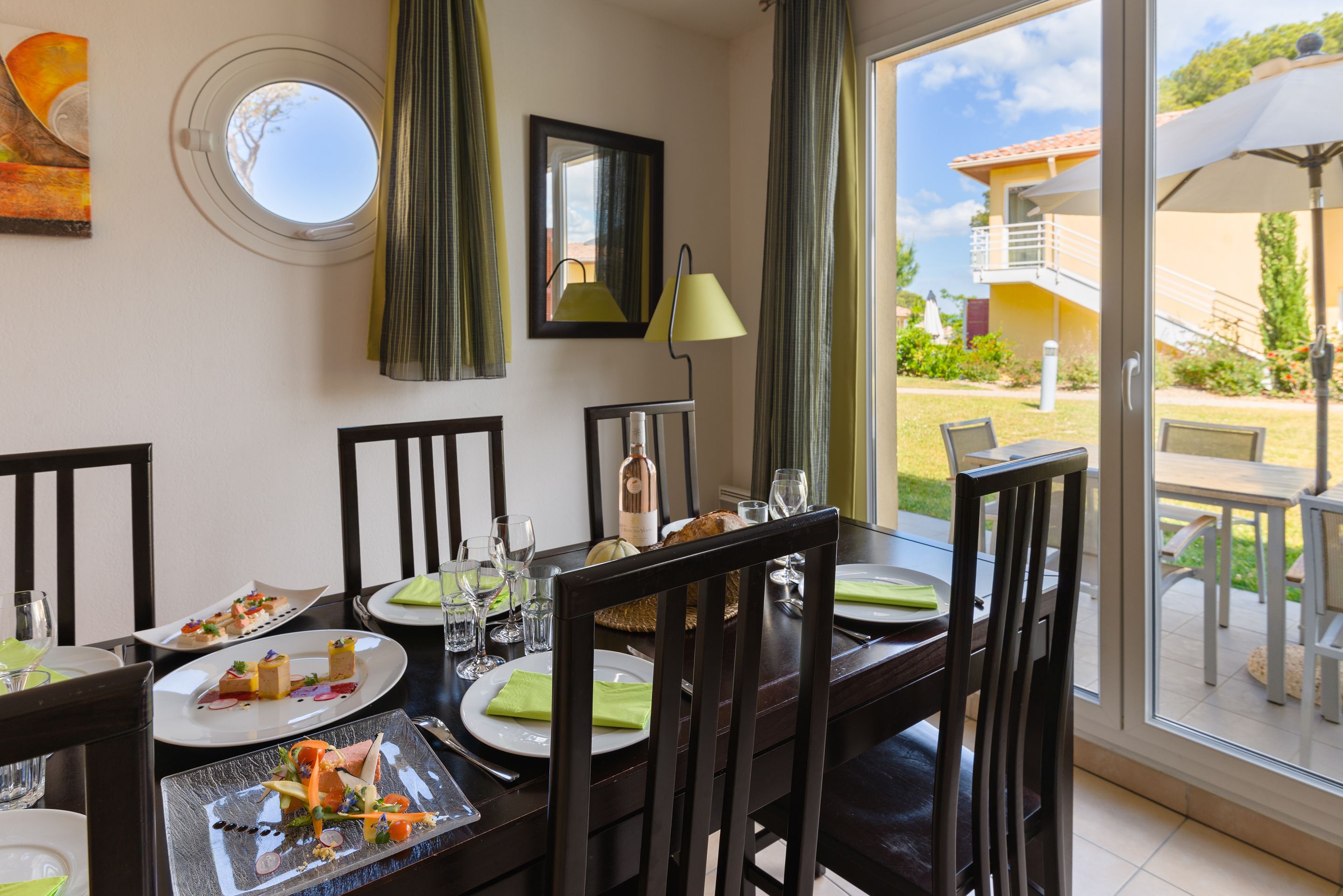 A dining area with a table set for four, chairs, a window with a view of the outside, and a lamp on the table.