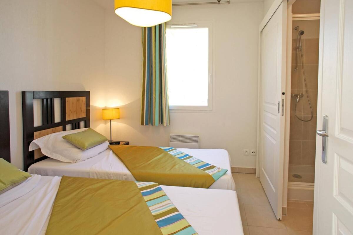 A hotel room with two beds, a window with curtains, a lamp, and a shower area.