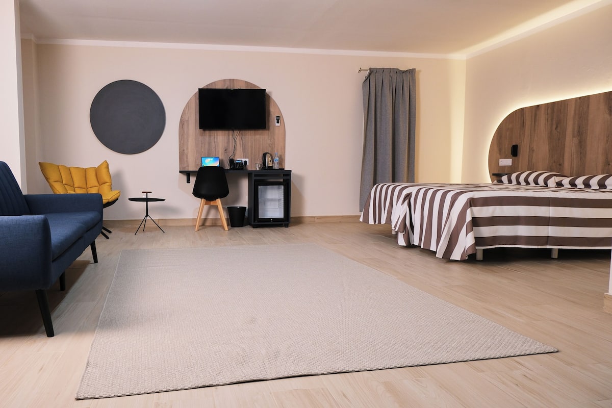 A modern hotel room with a blue sofa, a small desk with a TV, a bed with striped bedding, and a round wall decoration.