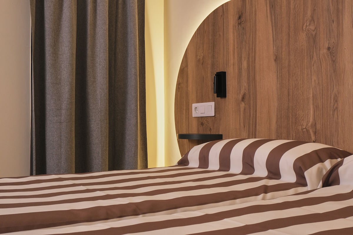 A hotel room with a wooden headboard, a bed with striped bedding, a nightstand, and a wall-mounted light switch.