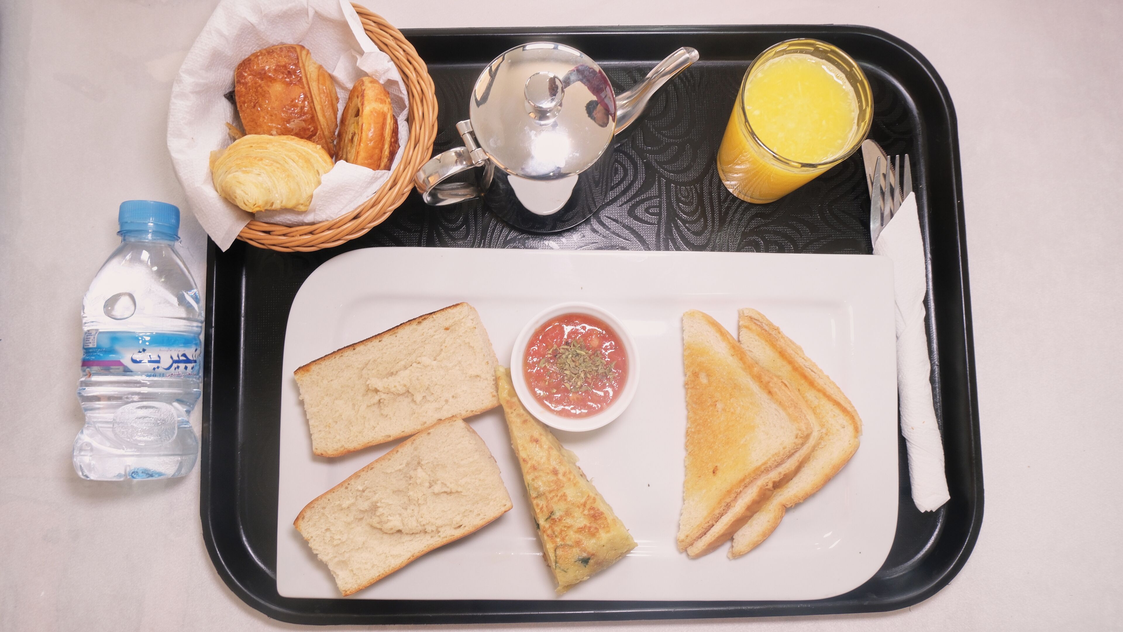 Una bandeja con tostadas, un cuenco pequeño de salsa, una botella de agua y una cesta de pasteles.