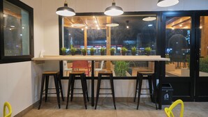 A restaurant interior with a long counter, multiple stools, hanging pendant lights, and a view of the street outside. - Hotel Aloe Emira NKC (Nouakchott)