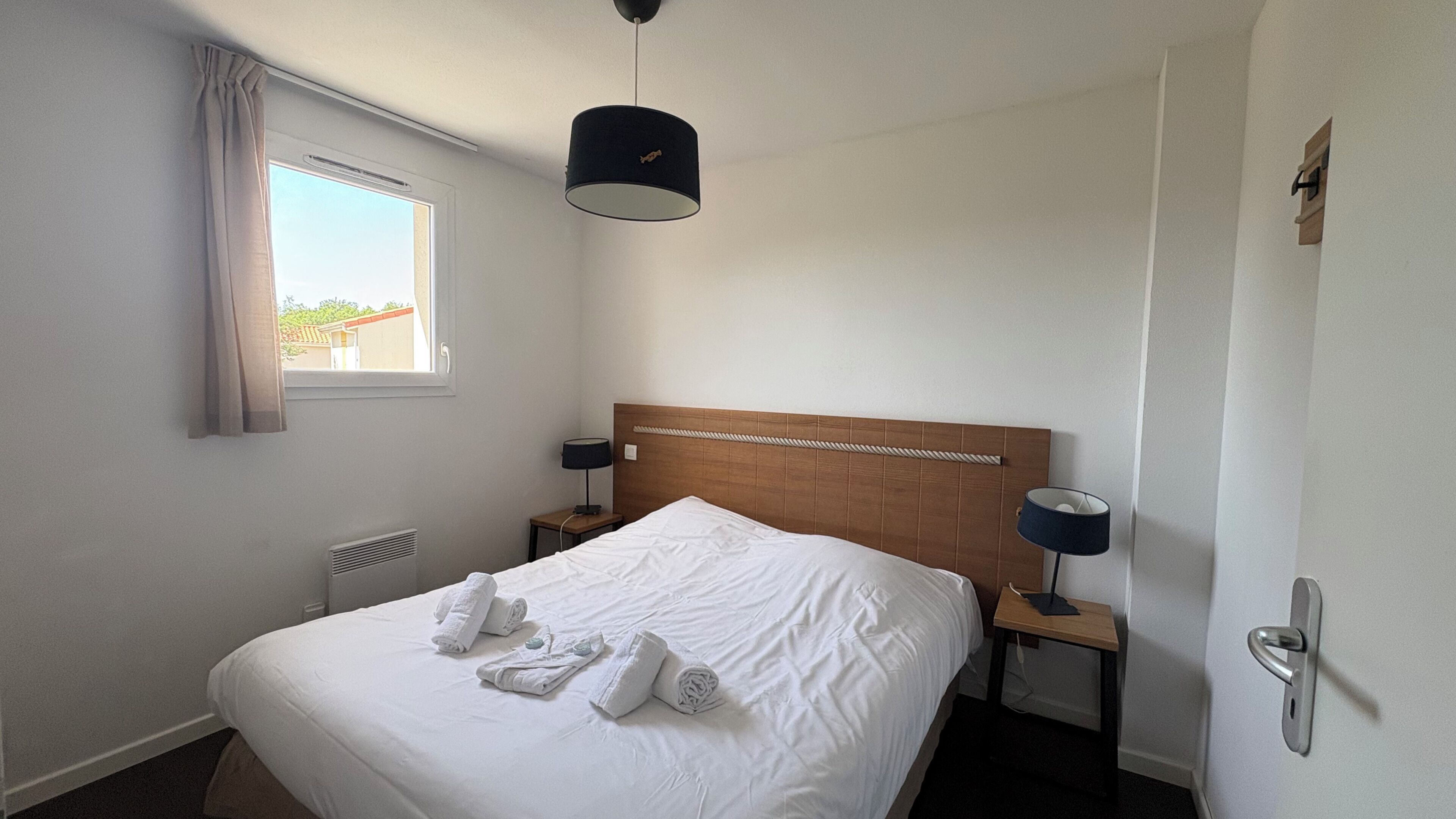 A hotel room with a bed, two bedside tables, a window with curtains, a ceiling light, and a door with a handle.