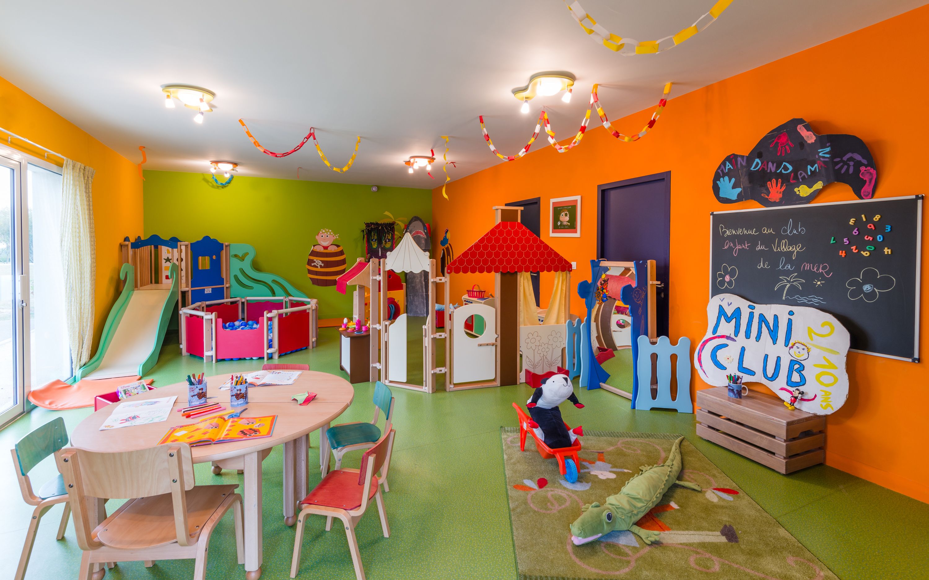 Una sala giochi vivace per bambini, con strutture colorate, una lavagna e un cartello con scritto "Mini Club".