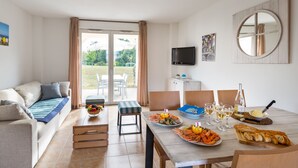 Una sala de estar moderna con un sofá, una mesa de comedor dispuesta con mariscos, copas de vino y pan. Un televisor está montado en la pared y hay un ventanal corredizo que da a un espacio exterior.