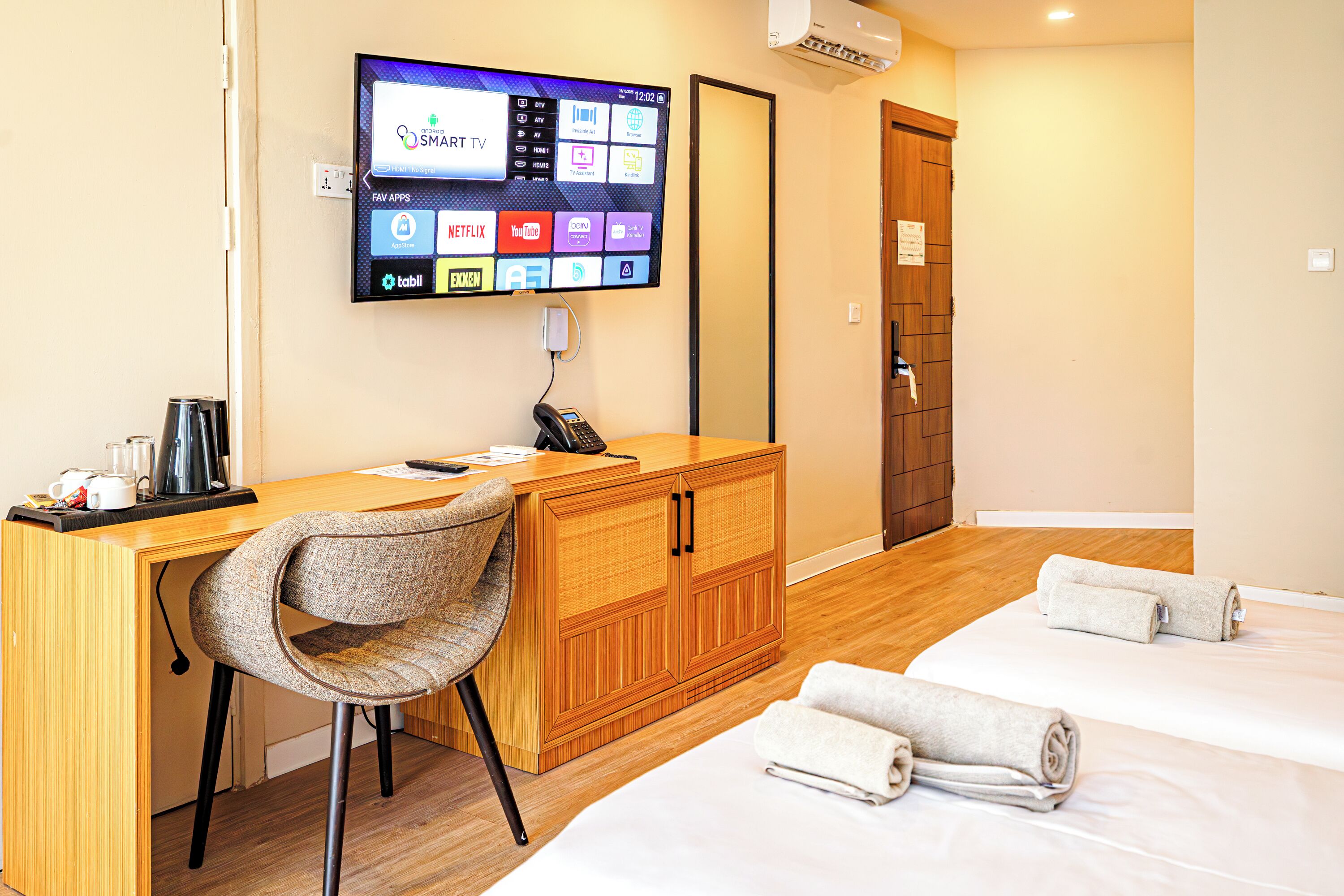 A hotel room with a wooden desk, a chair, a bed with towels, and a TV displaying various apps.