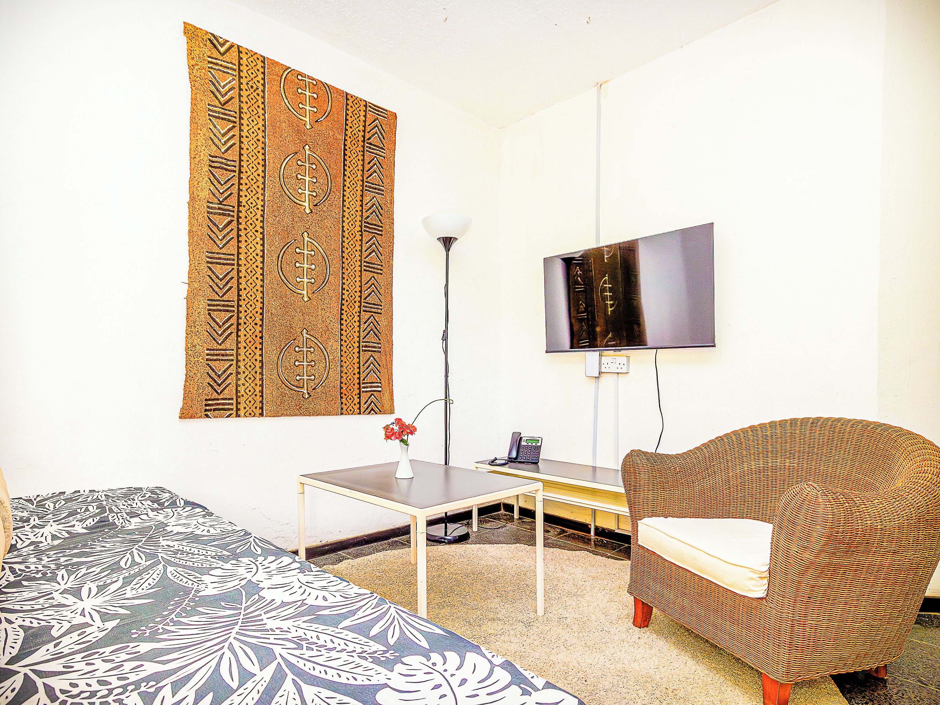 A room with a patterned wall hanging, a television mounted on the wall, a wicker chair, a small table with a vase of flowers, and a sofa with a floral-patterned cover.