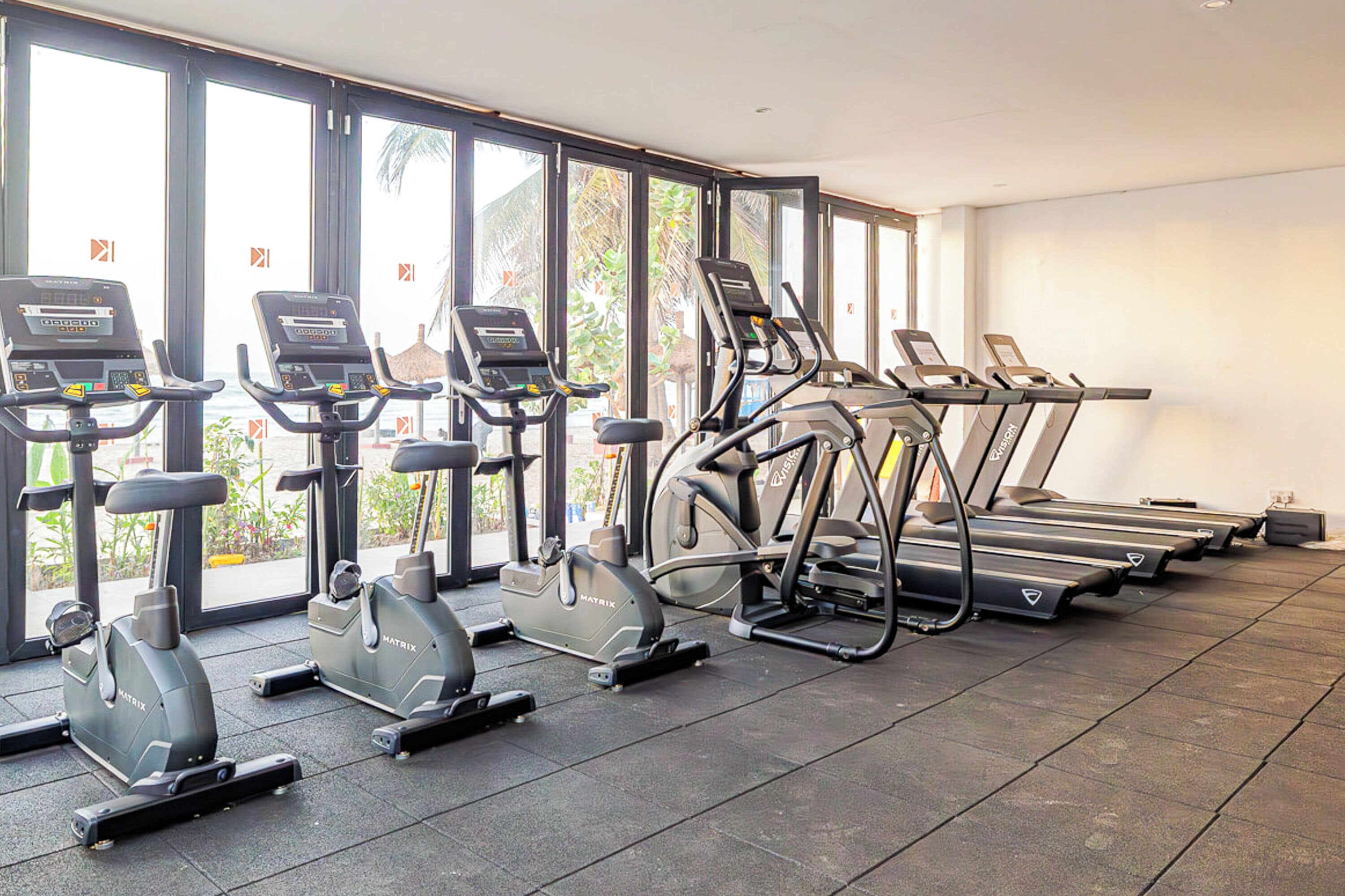 A gym with multiple treadmills and stationary bikes, large windows, and a tiled floor.
