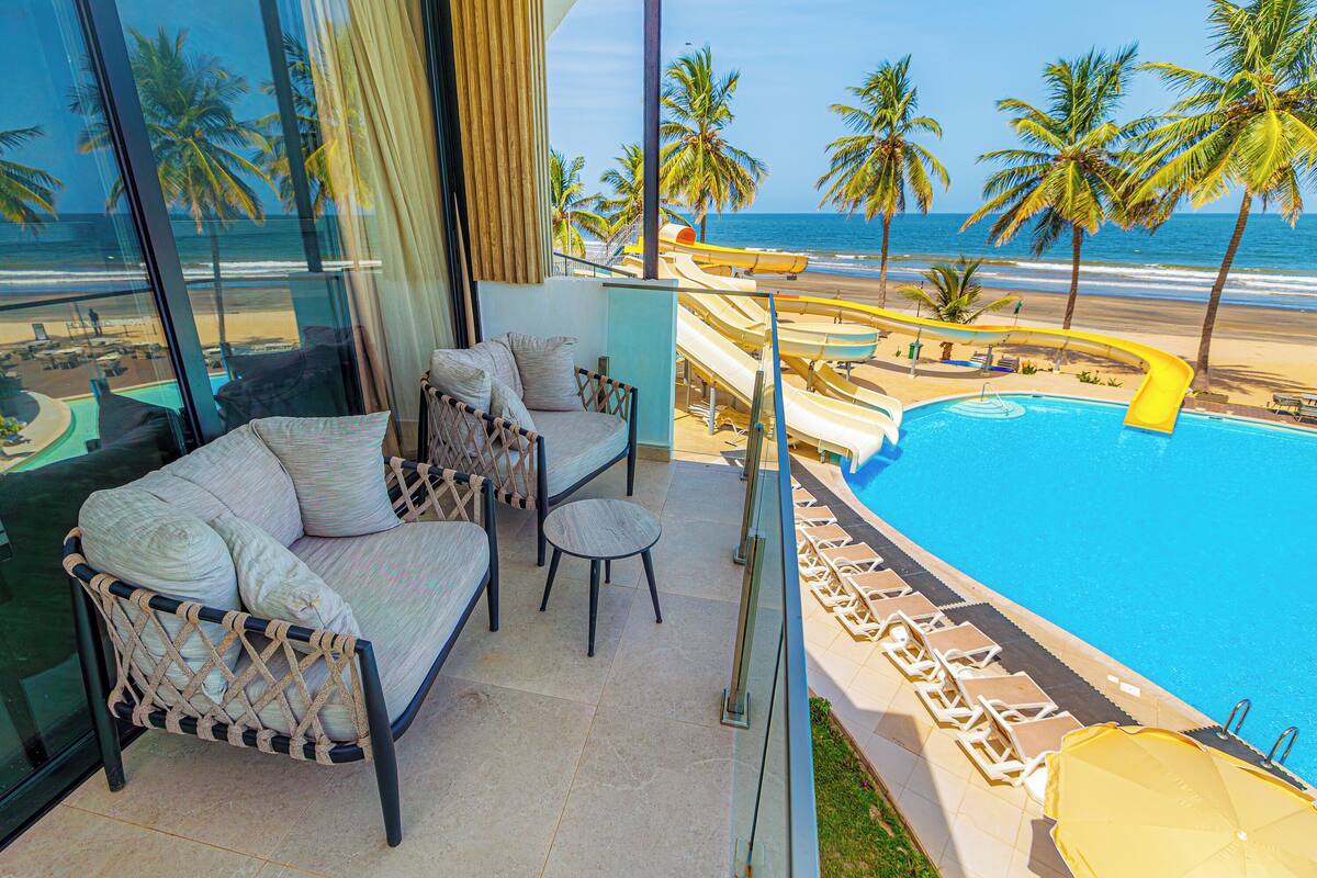 Un balcón con sofá, una mesita y una silla, con vista a una piscina y playa con palmeras.