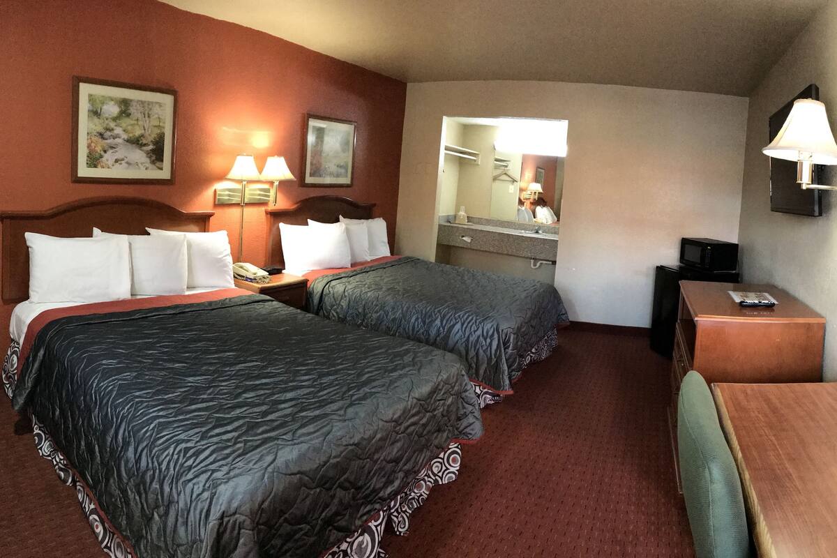 A hotel room with two beds, a desk, a chair, a dresser, a mirror, and two wall-mounted lamps.