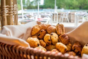 A basket of assorted pastries, wicker furniture, and outdoor seating with a view of a body of water. - Résidence Odalys Erromardie Plage (Saint-Jean-de-Luz)