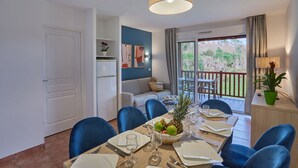 Un comedor con una mesa dispuesta para seis personas, una cocina al fondo y un balcón con vistas a árboles.
