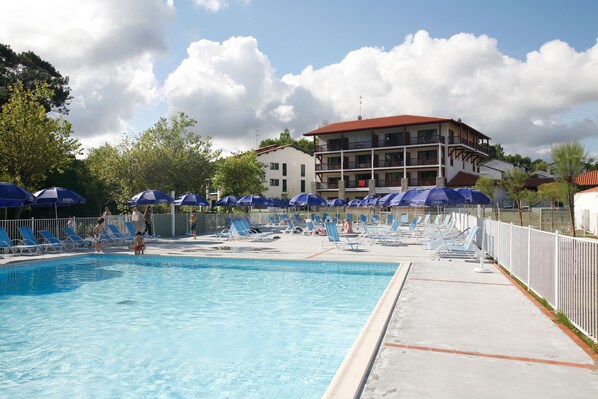 A swimming pool with lounge chairs and umbrellas, surrounded by a white fence and a building with balconies. - Résidence Odalys Erromardie Plage (Saint-Jean-de-Luz)