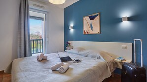 Una cama bien tendida con una laptop, un libro y una toalla enrollada. Hay una maleta y una lámpara a un lado.
