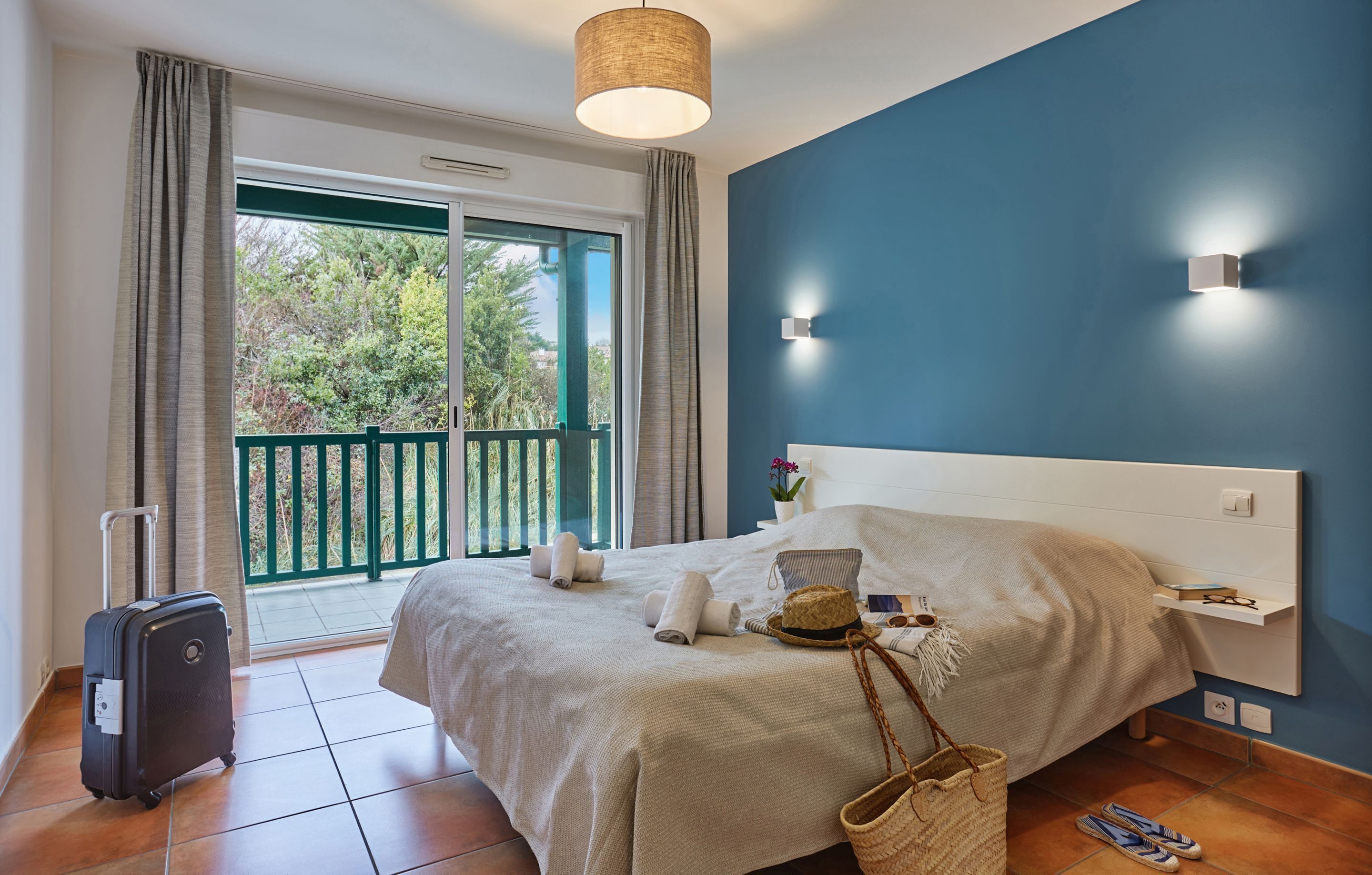 Ein Hotelzimmer mit Bett, Koffer, Balkon mit Blick auf Bäume und einer Wandleuchte.