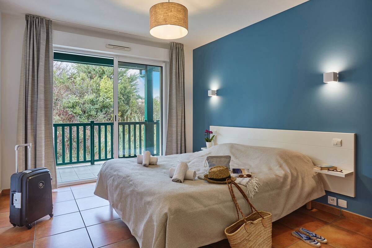 Ein Hotelzimmer mit Bett, Koffer, Balkon mit Blick auf Bäume und einer Wandleuchte.