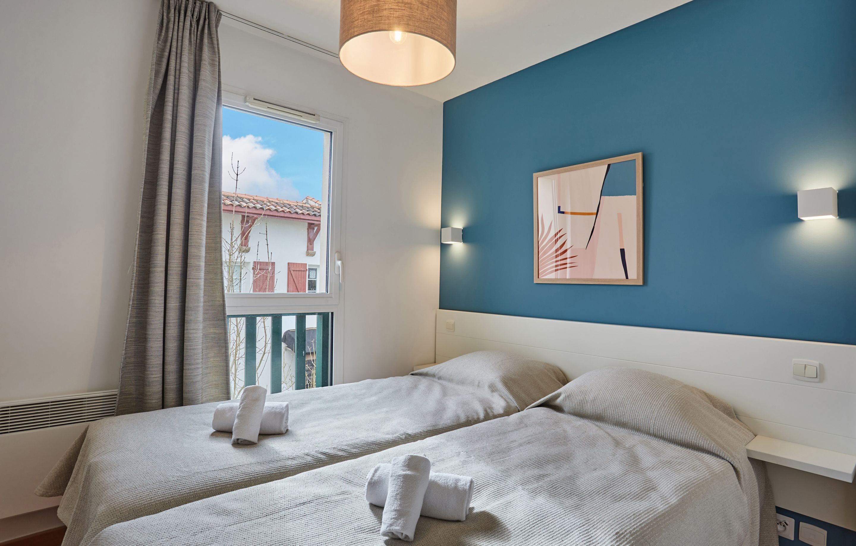 Ein Hotelzimmer mit zwei Betten, einem Fenster mit Vorhängen, einer blauen Akzentwand und einem gerahmten Bild.