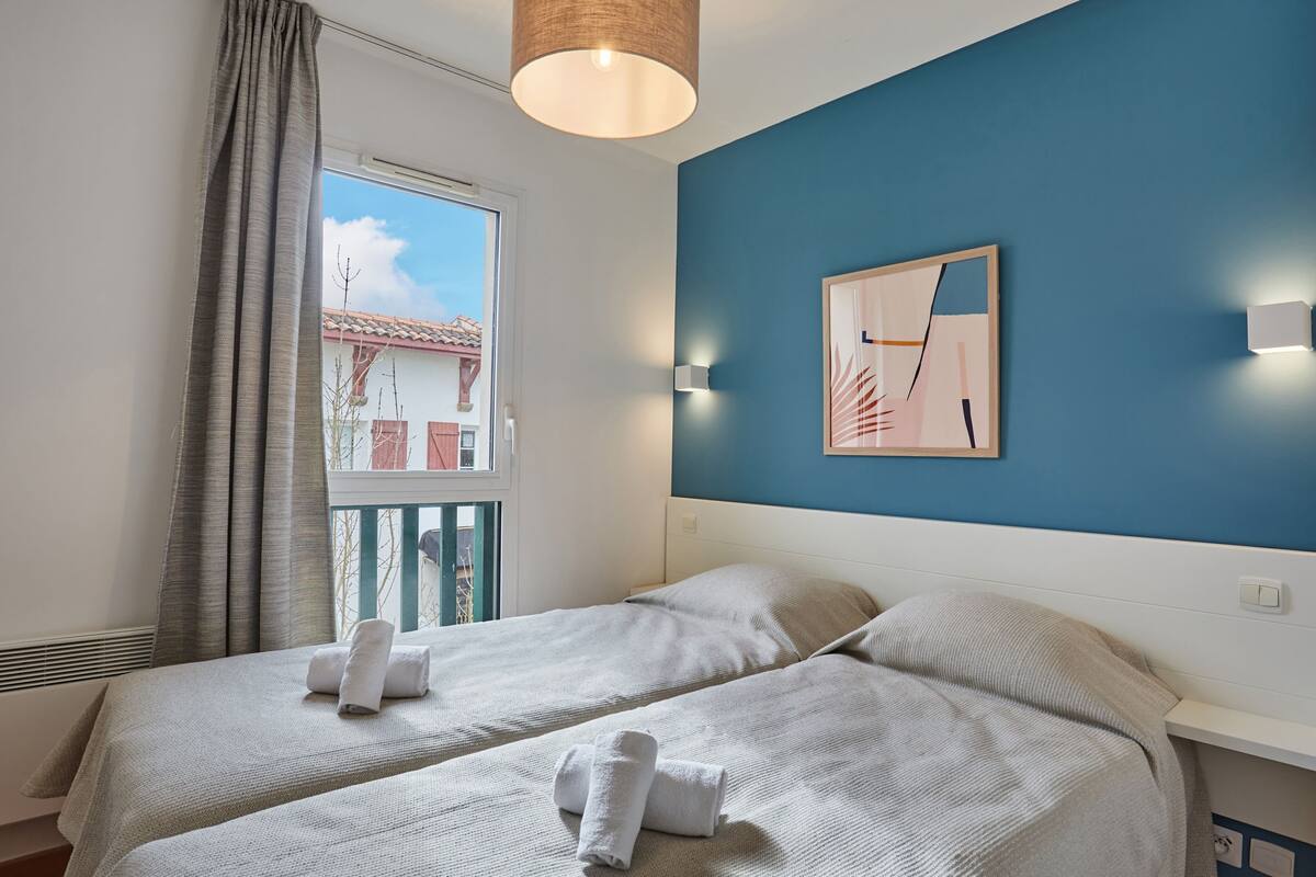 Ein Hotelzimmer mit zwei Betten, einem Fenster mit Vorhängen, einer blauen Akzentwand und einem gerahmten Bild.