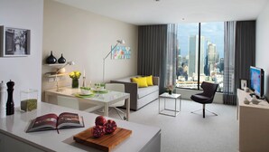 Uma sala de estar moderna com vista para a cidade, uma área de refeições com um livro e frutas, e um espaço de estar acolhedor com um sofá e uma poltrona.