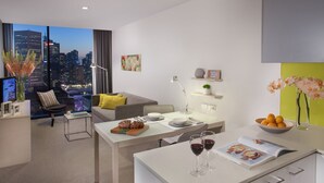 Uma sala de estar moderna e bem iluminada, com vista para a cidade, uma área de jantar com mesa posta para dois e um espaço de estar acolhedor com sofá e poltrona.