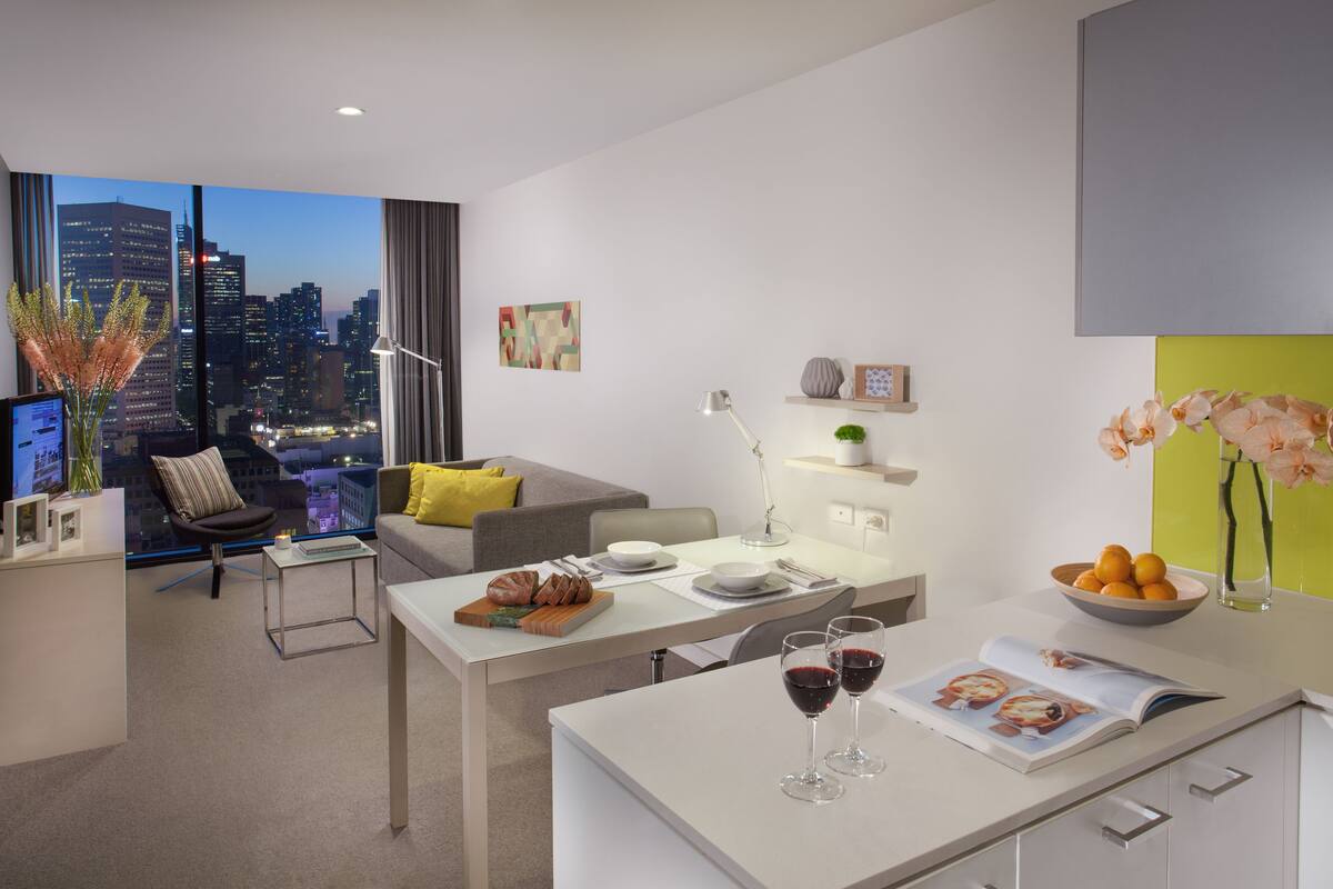 A modern, well-lit living room with a city view, a dining area with a table set for two, and a cozy seating area with a sofa and a chair.