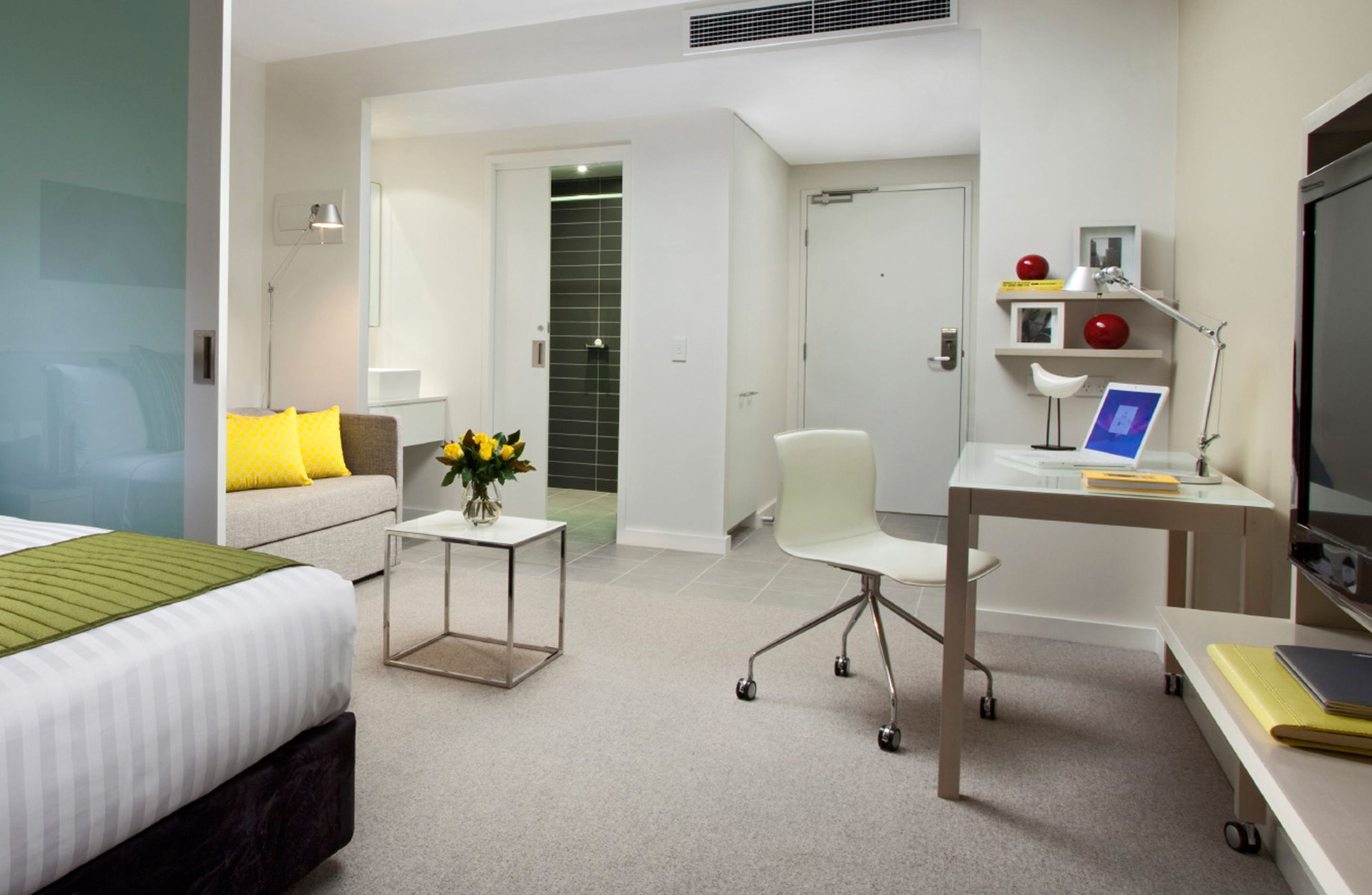 A modern hotel room with a bed, a desk, a chair, a small table with a vase, and a TV.