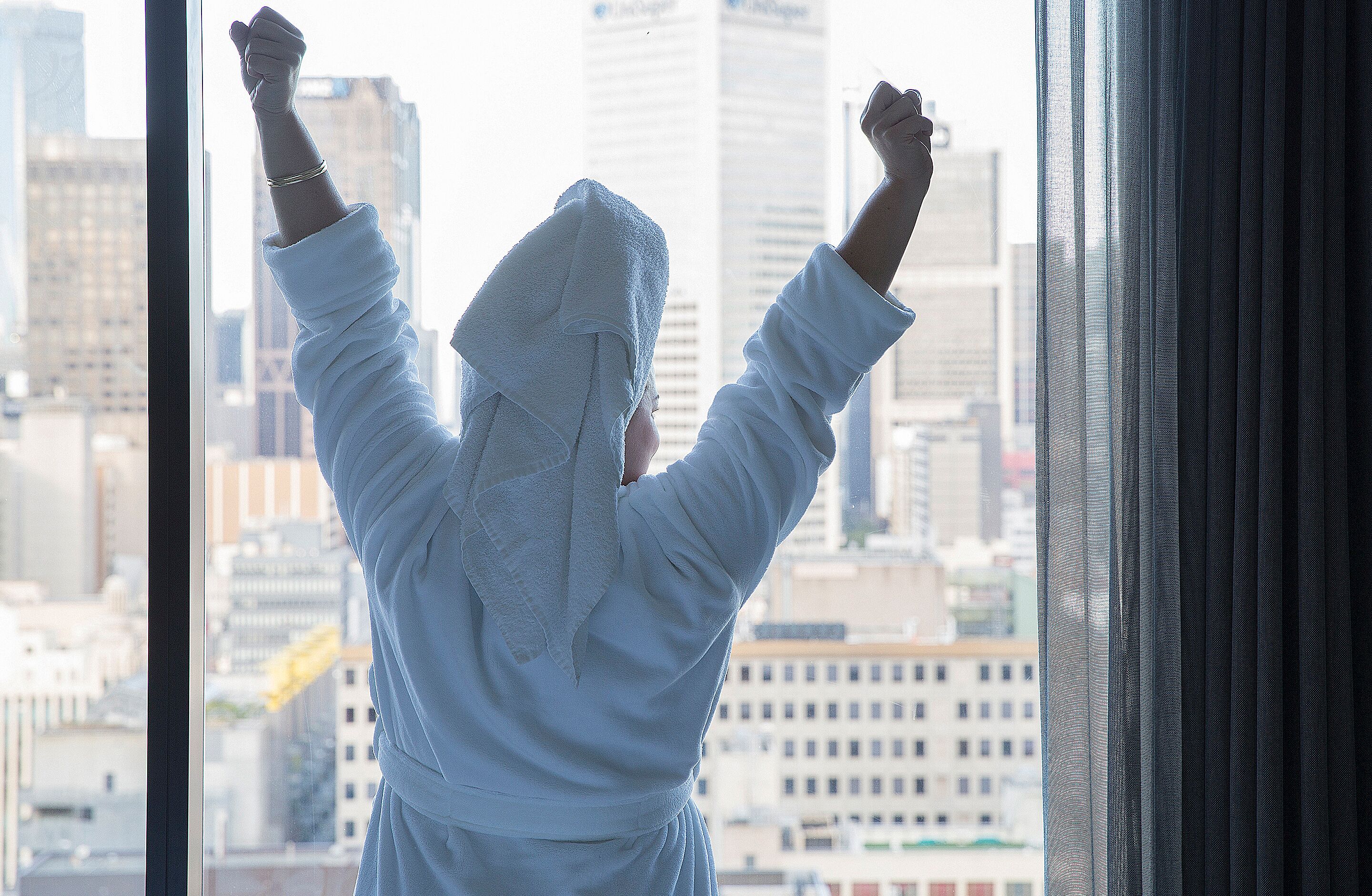 A person in a white robe with arms raised, looking out of a window with a cityscape view.