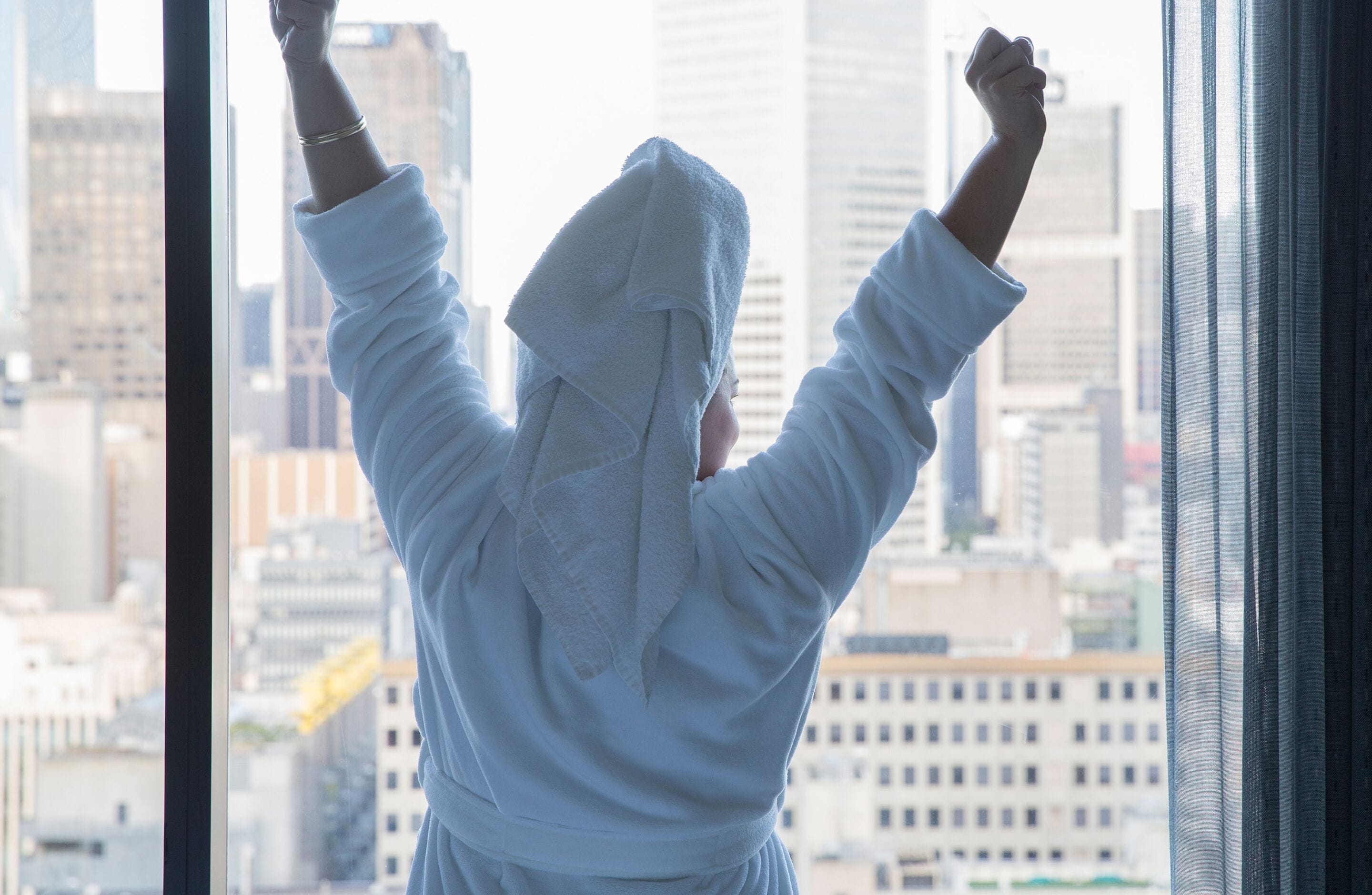 A person in a white robe standing by a window with a cityscape view.
