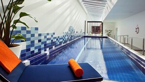 Uma piscina interior moderna com um revestimento de azulejos azuis, uma planta em vaso e uma espreguiçadeira com toalha laranja.