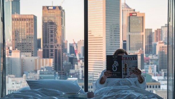 Uma pessoa lendo um livro num quarto com vista para a cidade pela janela.