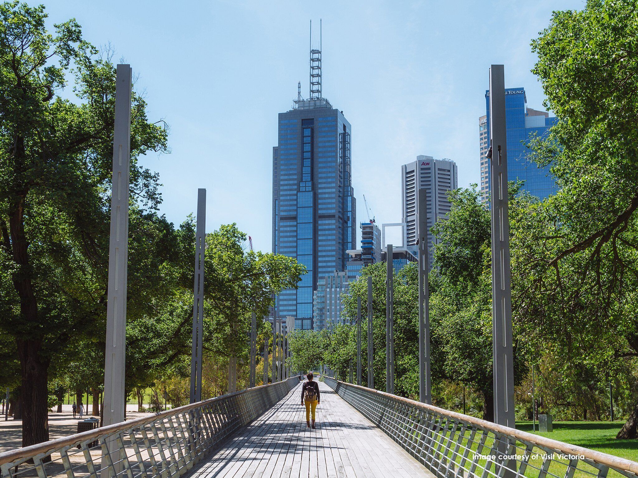 A pedestrian bridge with a metal railing, surrounded by trees and leading towards a cityscape with tall buildings.
