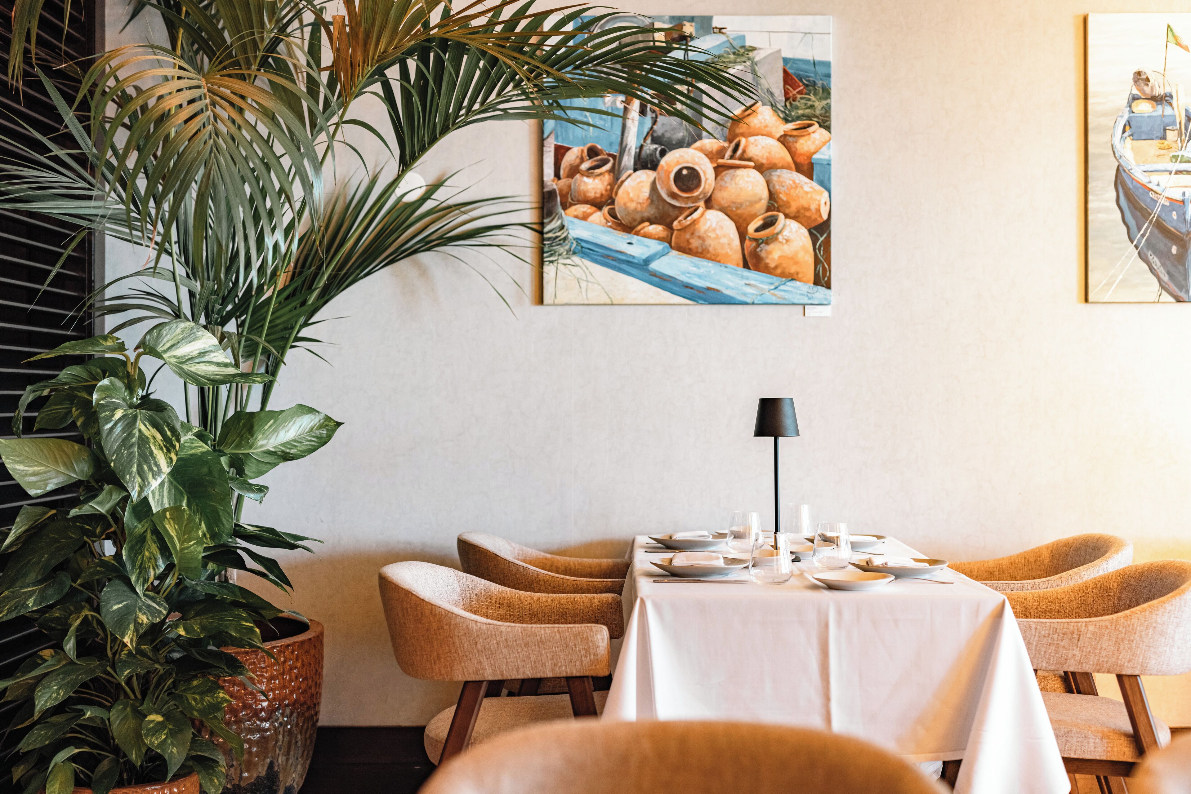A dining area with a table set for four, surrounded by four chairs, a large potted plant, and two paintings on the wall.