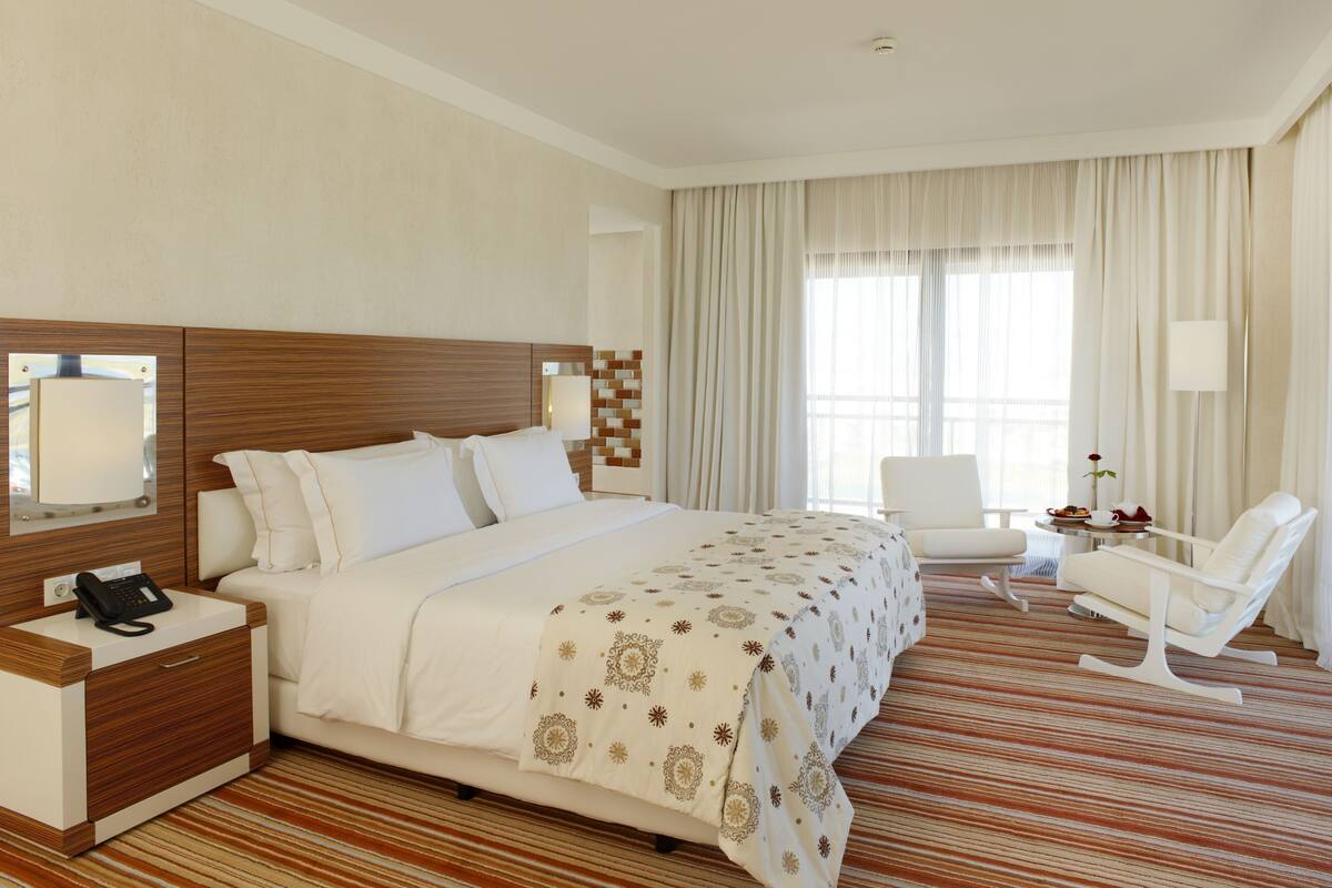 A hotel room with a large bed, bedside table with a phone, a chair, and a small table with a flower.