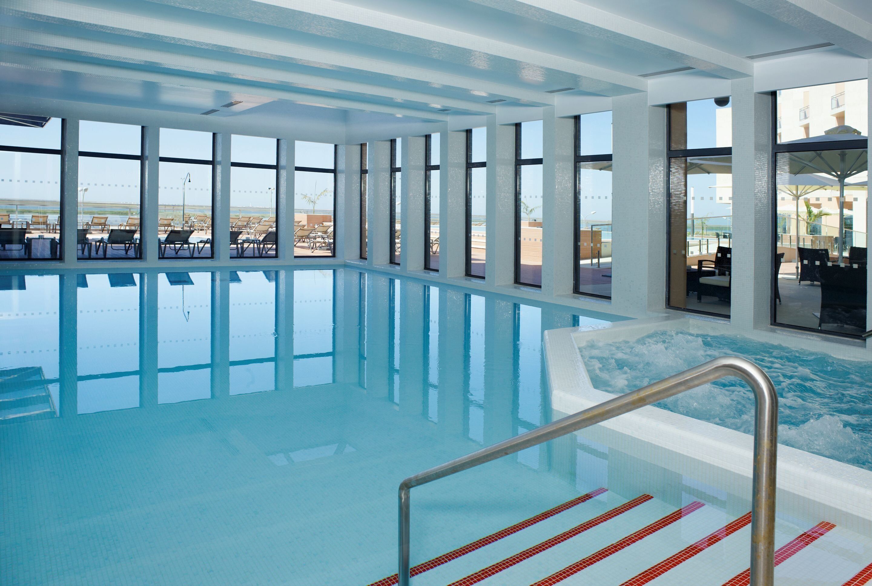 Piscina interior con agua azul cristalina, ventanas amplias y vista a la playa.