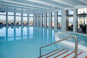 Piscina interior con agua azul cristalina, ventanas amplias y vista a la playa.