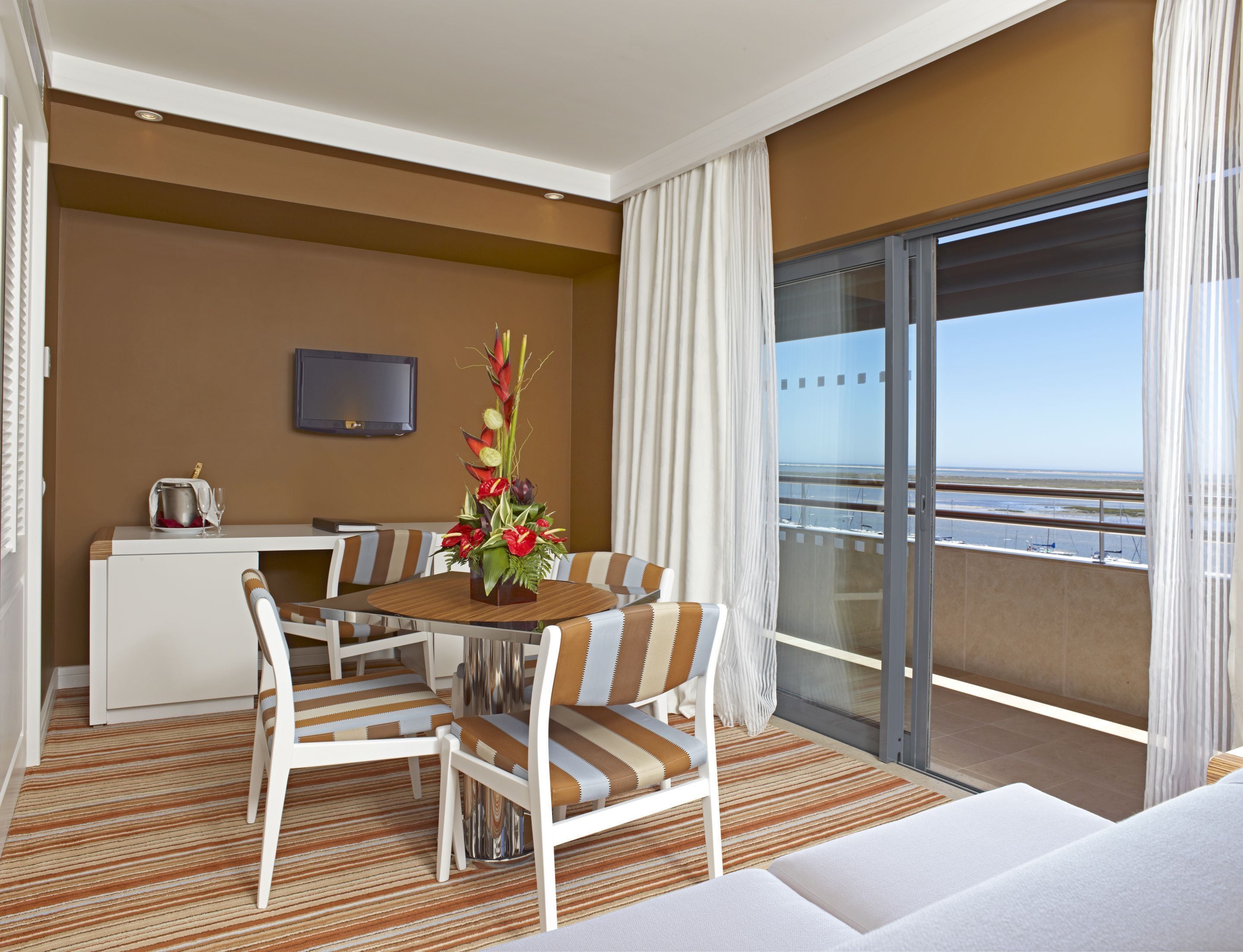 A hotel room with a dining table, chairs, a sofa, a TV, a balcony with a view, and a decorative plant.
