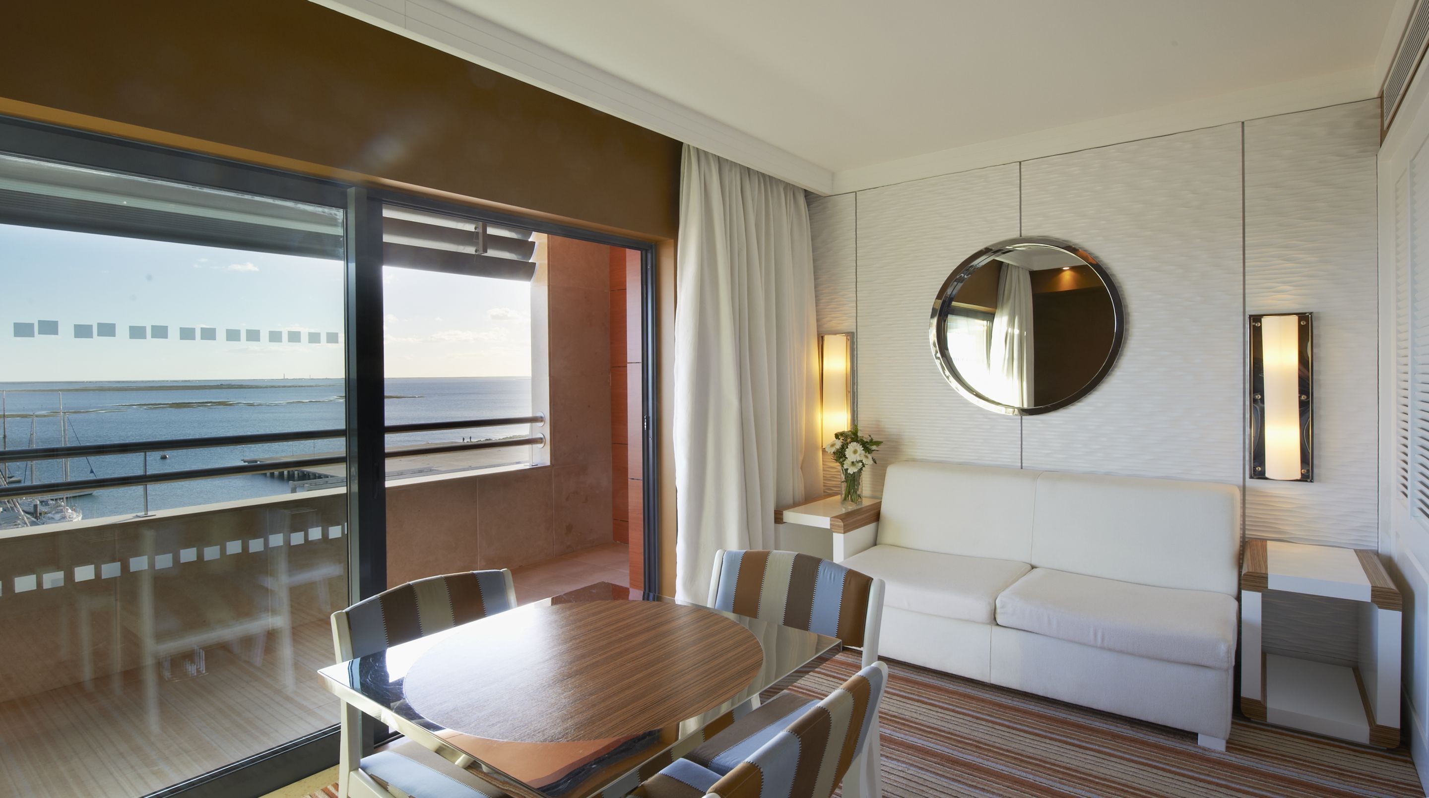 A hotel room with a large window offering a view of the sea, a round mirror on the wall, a white sofa, and a dining table with chairs.