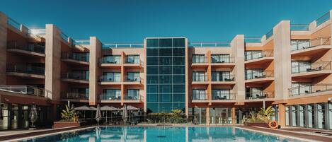 A modern hotel with a central glass facade, a swimming pool, and multiple balconies.