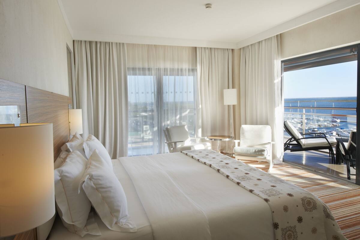 A hotel room with a large bed, a sitting area with a chair and table, and a balcony with a view of the marina.